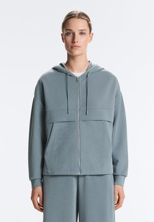 Nő világoskék bordázott, cipzáras kapucnis pulóverben, amelyen húzózsinórok vannak, és hozzáillő, bő fazonú nadrágot visel, sima fehér háttér előtt áll.