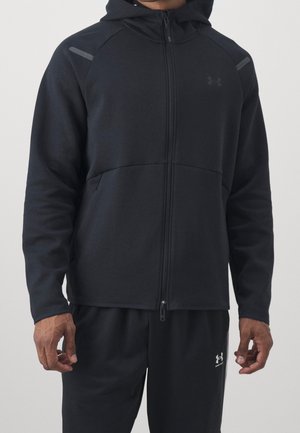 Mann trägt schwarzen Zip-Hoodie und schwarze Jogginghose mit kleinen Under Armour-Logos auf Brust und Bein, stehend vor einfarbigem Hintergrund.