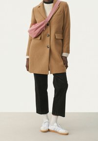 Manteau en mélange de laine de chameau avec des boutons noirs, associé à un pantalon en velours côtelé noir, des chaussures de sport blanches et un sac à bandoulière rose. Pull en tricot texturé en dessous.