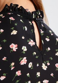 Black blouse with small floral pattern in pink and white. Features a ruffled neckline and a button closure at the back. Soft fabric texture.