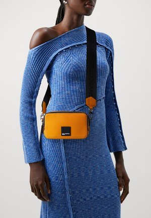 Sac bandoulière jaune avec une finition texturée, sangle noire avec logo, forme rectangulaire plate, assorti à une robe côtelée bleue.