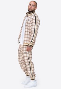 Beige karierter Trainingsanzug mit schwarzen und goldenen Streifen, bestehend aus einer Zip-Jacke und elastischen Bündchen, kombiniert mit weißen Turnschuhen.