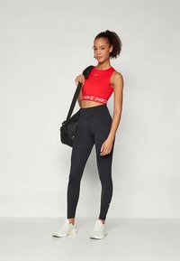 Rdeč Nike Pro kratki top z belim logotipom, visok pas črne legice in bele superge. Manekenka nosi črno torbo čez ramo.