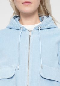 Ανοιχτό μπλε φούτερ με φερμουάρ από κορντόροι, με κουκούλα, ασημένιο φερμουάρ και δύο μπροστινές τσέπες, που διαθέτει ριγωτή υφή και κορδόνια στο τελείωμα.