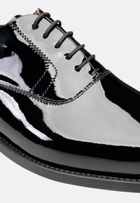 Scarpa in pelle verniciata nera con finitura lucida, dotata di sei occhielli, lacci neri e cuciture dettagliate lungo i bordi.