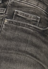 Lähellä mustat farkut, osoittaen takataskua, jossa on näkyvät tikit ja metallinen niitti teksturoituneessa kankaassa.