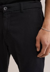 Fekete pamut nadrág slim fit szabással, lapos elülső résszel, gombos záródással és sima textúrával. Egy kéz a comb területén pihen.