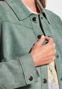 Groene suede jas met een klassieke kraag, zwarte drukknoopknopen en een gedetailleerde bloemenvoering die zichtbaar is bij de opening.