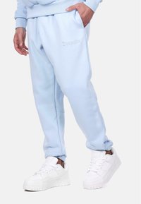 Light blue sweatpants made of soft fabric, featuring an embroidered logo and side pockets, paired with white sneakers.