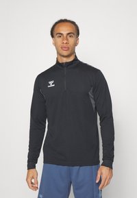Top sportivo zip-up nero con maniche lunghe, presenta dettagli grigi sui lati e un logo sul petto. Texture liscia e design aderente.