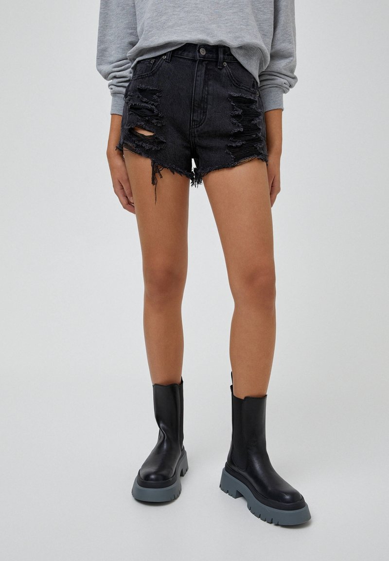Personne portant un short en jean noir déchiré, un haut gris à manches longues et des bottes noires à plateforme épaisses, debout sur un fond gris clair.