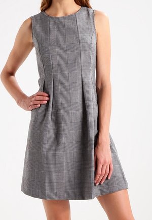 Robe sans manches à carreaux gris avec des plis à la taille, portée par une personne avec une main sur la hanche et l'autre reposant le long du corps.