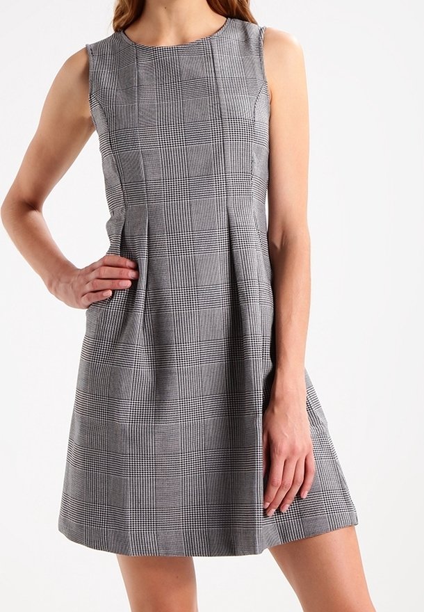 Robe sans manches à carreaux gris avec des plis à la taille, portée par une personne avec une main sur la hanche et l'autre reposant le long du corps.