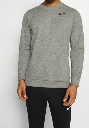 Mand iført en grå langærmet Nike sweatshirt og sorte Nike bukser, stående mod en ensfarvet baggrund.
