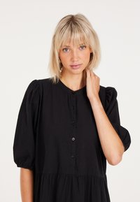 Musta nappipaita mekko, jossa on puffihihat ja kerroksellinen hame. Valmistettu teksturoidusta kankaasta, jossa on pyöreä pääntie ja mustat napit.