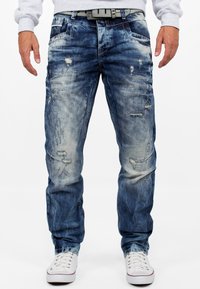 Jeans in denim blu sbiadito con dettagli strappati, taglio dritto, cuciture visibili e tasche; abbinati a sneakers bianche.