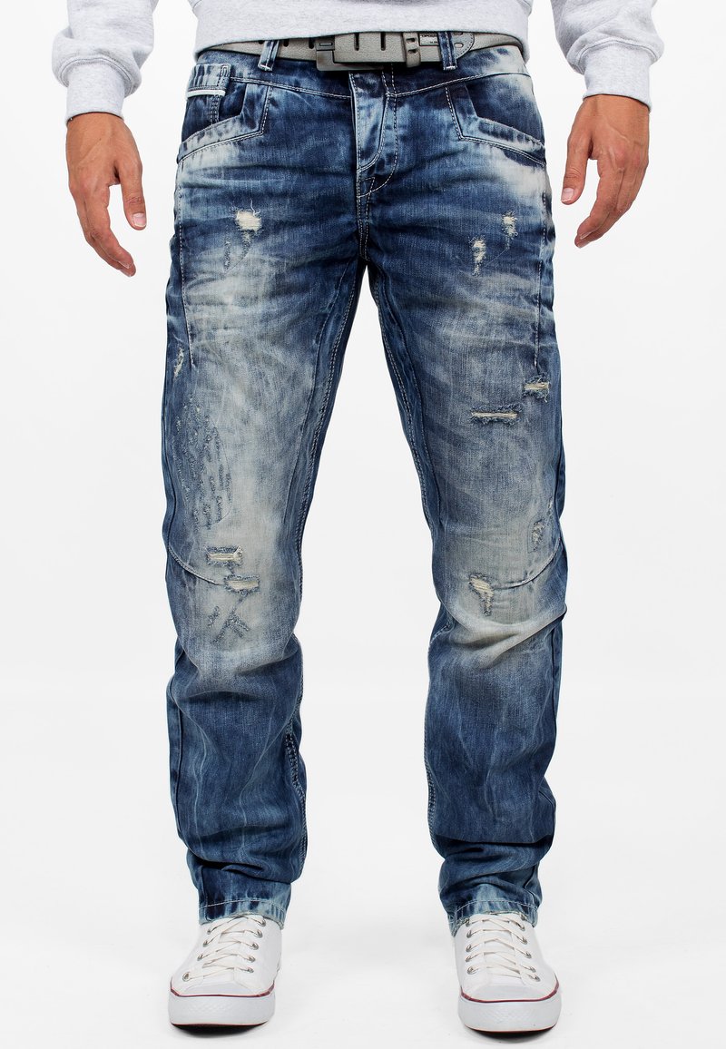 Jeans in denim blu sbiadito con dettagli strappati, taglio dritto, cuciture visibili e tasche; abbinati a sneakers bianche.