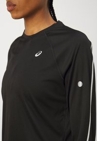 Černé sportovní tričko s dlouhým rukávem a kulatým výstřihem, s malým bílým logem na hrudi a bílým akcentem na rukávu.
