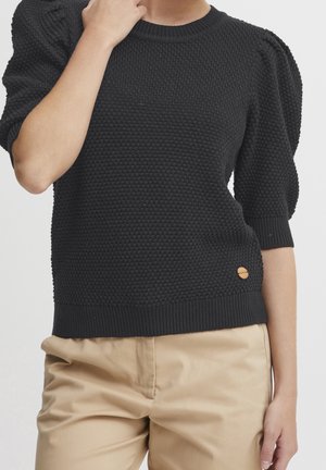 Personne portant un pull en tricot noir texturé avec des manches mi-longues bouffantes et un pantalon beige, la main touchant le cou, sur un fond uni.