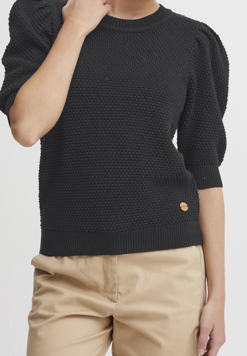 Personne portant un pull en tricot noir texturé avec des manches mi-longues bouffantes et un pantalon beige, la main touchant le cou, sur un fond uni.
