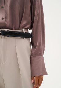 Satin, mauve långärmad skjorta i kombination med beige byxor, fäst med ett svart läderbälte. Skjortan har en knapp-ned design och manschetter.