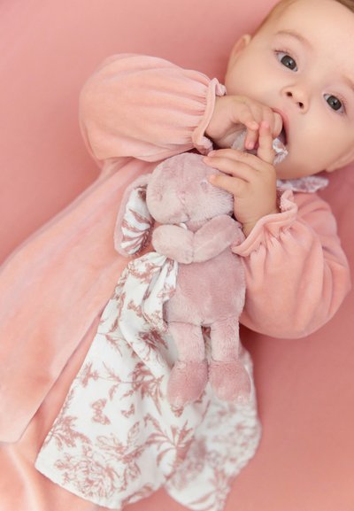Rosa plyschonesie med volangärmar, håller en ljusrosa kaninleksak och ett vitt tyg med rosa blommönster, mot en mjuk rosa bakgrund.