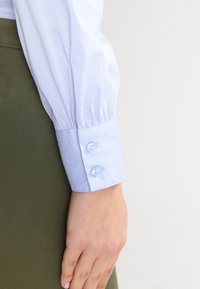 Ljusblå skjorta med volangärmar och knappdetaljer, som bärs med en grön kjol. Tyget ser slätt och polerat ut.