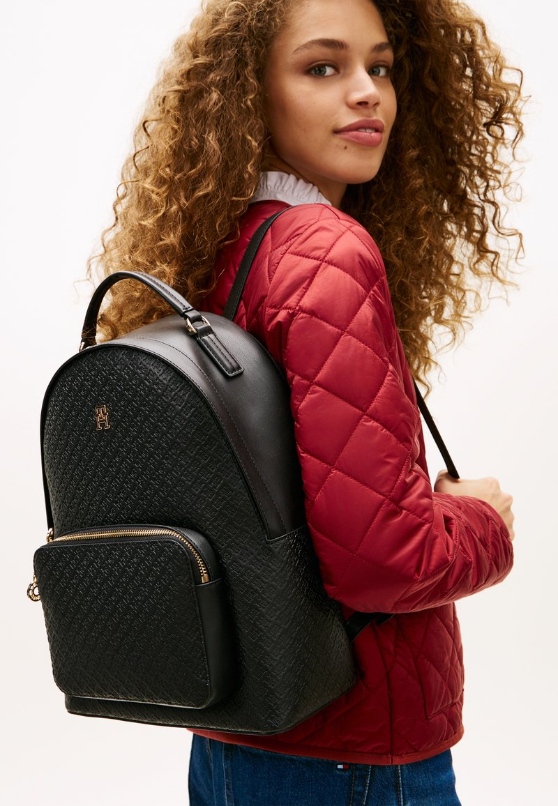 Νεαρή γυναίκα με σγουρά μαλλιά που φοράει κόκκινο μπουφάν παγωτό και κρατάει μια υφή μαύρη τσάντα πλάτης με χρυσές λεπτομέρειες φερμουάρ.