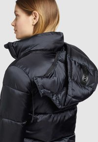 Giovane donna di profilo che indossa un piumino nero lucido con cappuccio parzialmente staccato su uno sfondo grigio uniforme.