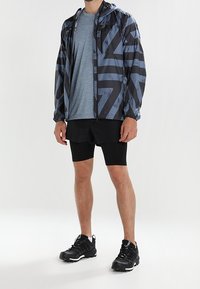Veste légère noire et grise à motif, fabriquée en matériau résistant à l'eau, portée avec une chemise bleue et un short noir, accompagnée de chaussures de sport noires.