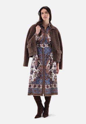 Frau trägt ein langes geblümtes Kleid mit Gürtel, eine braune Wolljacke über die Schultern gelegt und kniehohe dunkelbraune Stiefel.