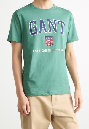 Grøn bomulds t-shirt med "GANT" i store blå bogstaver, et rødt skjoldlogo og "AMERICAN SPORTSWEAR" i hvid tekst nedenfor.