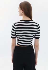 Schwarz-weiß gestreifter, kurzes Sweatshirt mit kurzen Ärmeln, das einen strukturierten Stoff und einen runden Ausschnitt zeigt. Getragen mit schwarzen Hosen.