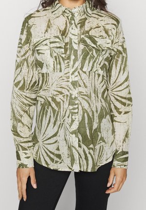 Femme portant une chemise boutonnée à motif feuilles de palmier vert et blanc, avec deux poches sur la poitrine et des manches longues.