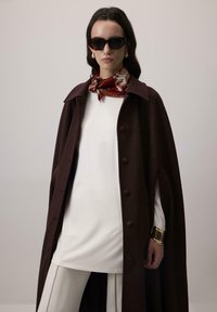 Sieviete tērpta tumši brūnā pogātā apmetnī, baltā apģērbā, ar sarkanu rakstainu šalli, lielu zelta aproci un tumšiem saulesbrillēm uz vienkārša fona.