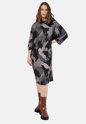 Langärmliges Kleid in Schwarz, Grau und Weiß mit abstraktem Muster, aus weichem Stoff. Kombiniert mit braunen Stiefeln mit Absatz und dicken Sohlen.