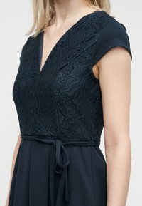 Γυναίκα που φοράει φόρεμα σε ναυτικό μπλε χρώμα με δαντελένιο μπούστο σε σχήμα V, κοντά μανίκια και ζώνη δεμένη στη μέση.