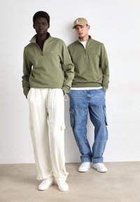 Twee modellen staan naast elkaar: de een draagt een groene zip-up sweatshirt en crèmekleurige broek; de ander heeft een groene sweatshirt, jeans en een pet aan.