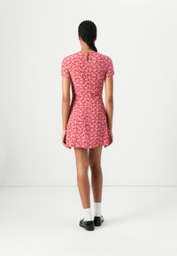 Robe fleurie rouge à manches courtes, corps ajusté et jupe évasée. Présente un détail de dos en forme de clé. Portée avec des chaussures noires et des chaussettes blanches.