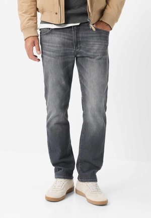 Uomo che indossa jeans grigi sbiaditi, giacca beige, maglione grigio, scarpe da ginnastica bianche con suola in gomma, in piedi su sfondo bianco uniforme.