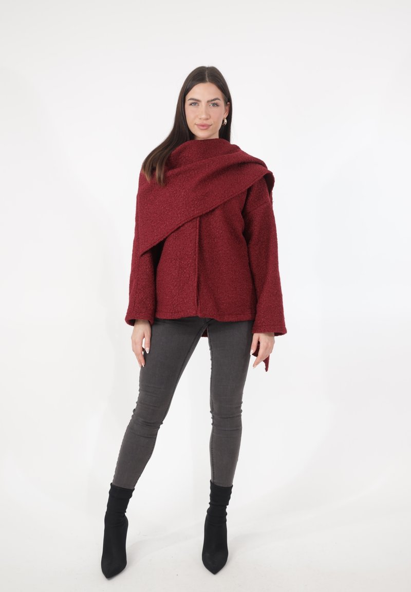 Giacca oversize color bordeaux con trama e design avvolgente. Abbinata a jeans grigi aderenti e stivaletti neri alla caviglia. Sfondo semplice.