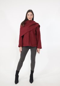 Bordeauxrode, oversized jas met textuur en een overslagontwerp. Gecombineerd met grijze slim-fit jeans en zwarte enkellaarsjes. Eenvoudige achtergrond.