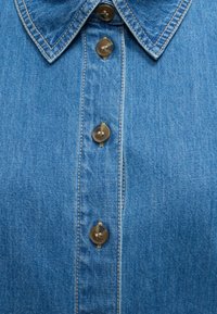 Κοντινή εικόνα μπροστινής όψης μπλε denim πουκάμισου που δείχνει γιακά, κάθετες ραφές και τέσσερα καφέ κουμπιά, κλεισμένα στο κέντρο.