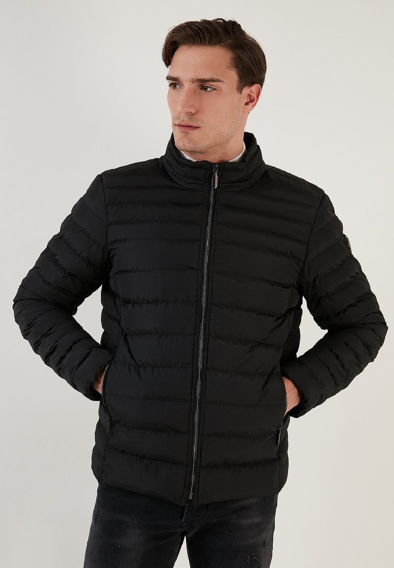 Jachetă puffer neagră cu guler înalt, design matlasat, închidere cu fermoar și buzunare laterale. Textură netedă cu detalii minimale de coasere.