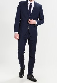 Completo blu navy realizzato in tessuto testurizzato, giacca aderente con un bottone, abbinato a pantaloni su misura e scarpe in pelle nera.