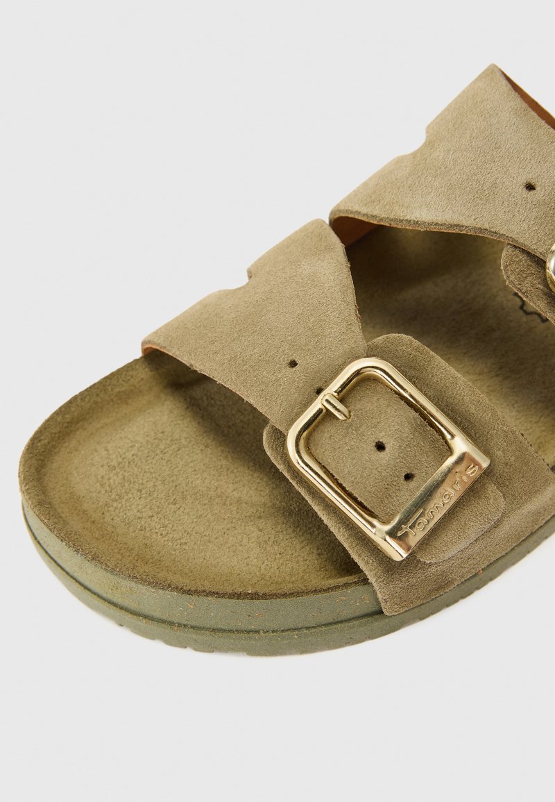 Nära bild av en taupe mockasandal med två remmar och ett gyllene spänne, med en texturerad fotbädd och en lätt böjd sula.