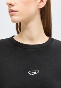 Νεαρή γυναίκα με ίσια καστανά μαλλιά, που φοράει μαύρο φούτερ με στρογγυλή λαιμόκοψη, το οποίο έχει ένα μικρό λευκό γεωμετρικό λογότυπο στο στήθος.