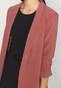 Blazer rosa-rossastro con un design strutturato, apertura frontale e maniche arrotolate; indossato sopra un top nero aderente. Texture liscia, dettagli minimi.