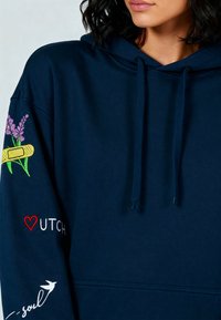 Marineblauwe hoodie met bloemenborduursel op de mouwen, trekkoorden en tekstaccenten. Zachte stof, casual ontwerp.