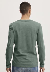 Grünes Langarmshirt mit lässiger Passform, rundem Halsausschnitt und weichem Material; getragen mit blauen Jeans.